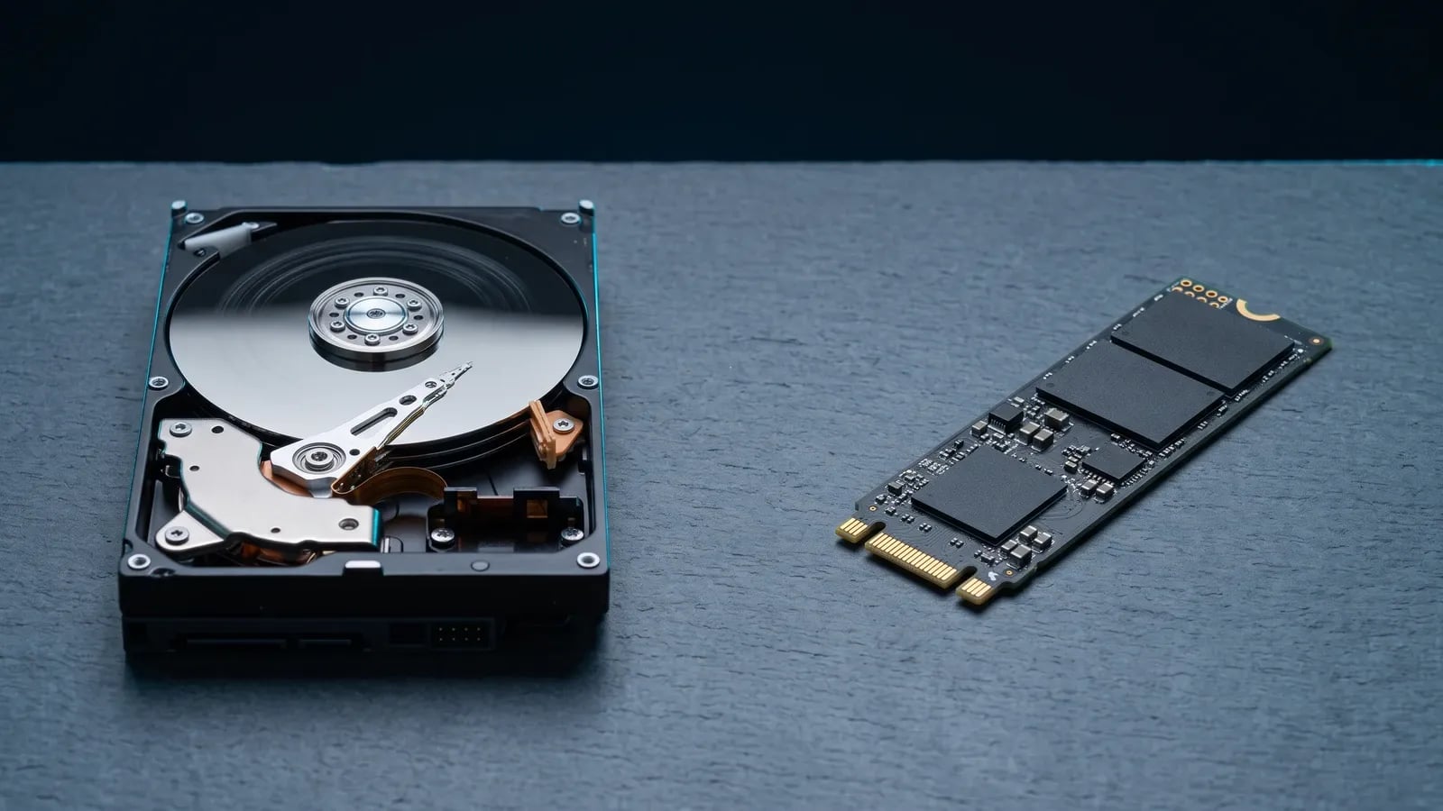 Un disque dur mécanique 3,5 pouces ouvert sur son plateau, à côté d'un SSD M.2 NVMe, sur une surface d'ardoise sombre