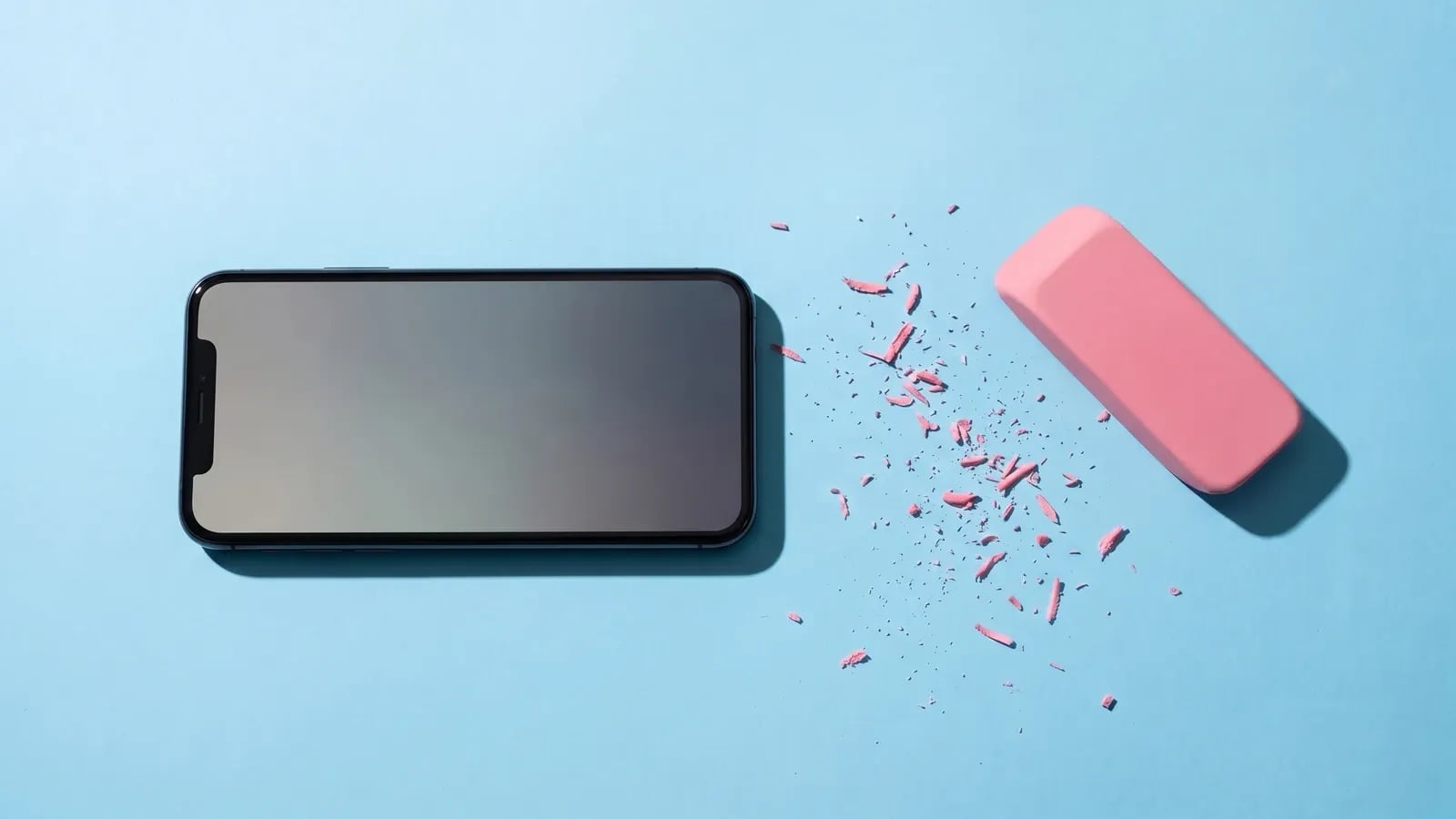 Smartphone posé à plat sur fond bleu ciel à côté d'une grande gomme rose et de copeaux, comme si son contenu venait d'être effacé